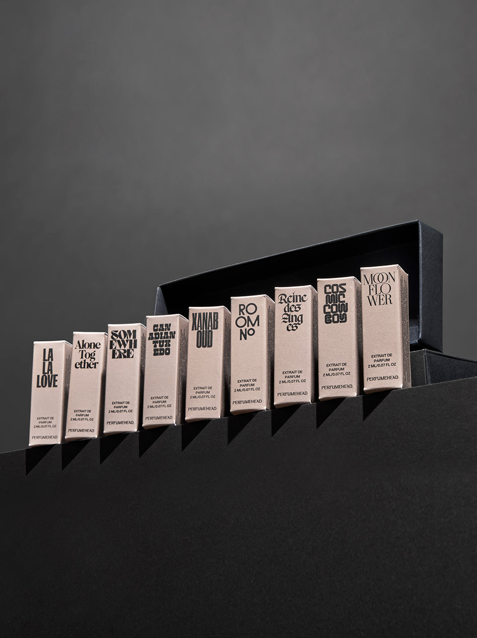 Nine slim rectangular perfume bottle boxes with cream-colored labels and black text are lined up on a dark surface against a dark gray background.