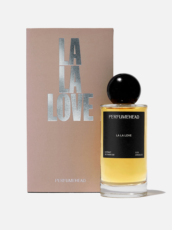 A bottle of Perfumehead 'LA LA LOVE' perfume with golden-yellow liquid and a black spherical cap, displayed next to its beige packaging box with metallic silver lettering, on a clean white background.