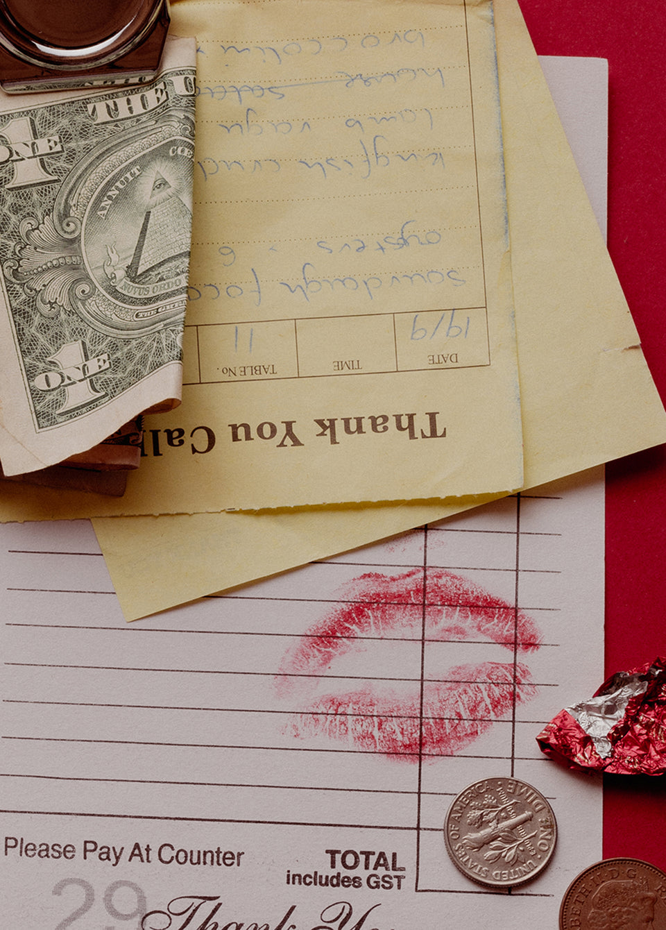 A styled flat-lay of vintage ephemera, featuring a yellow handwritten receipt, a red lipstick kiss mark on a white payment receipt, a US one-dollar bill, coins, and a piece of red foil against a red background.