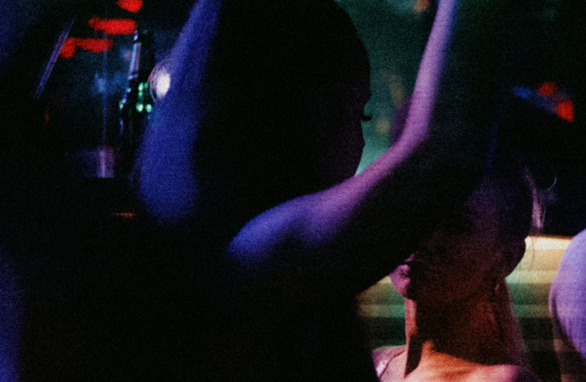 A dark, grainy, low-light photograph of a person with arms raised in a dimly lit environment, heavily tinted with blue and purple hues.