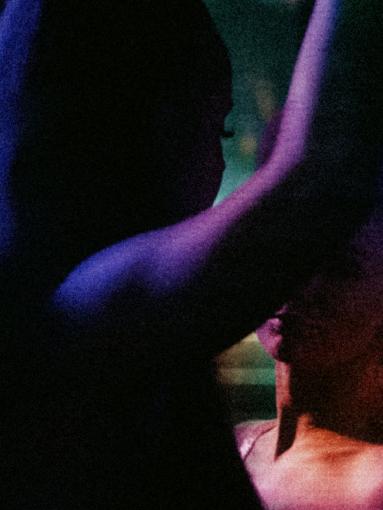 A close-up, grainy photograph of two people dancing under dramatic purple and blue lighting, with dark shadows obscuring much of the frame.