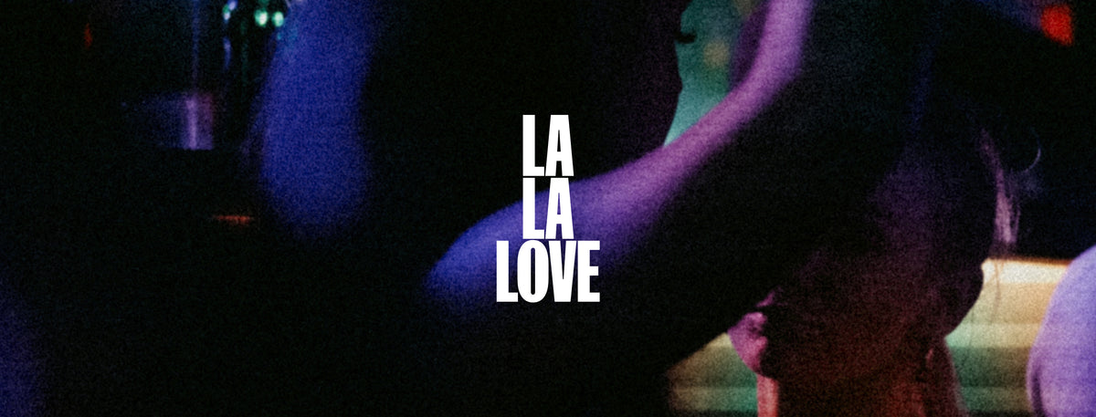 A dark, grainy, low-light photograph featuring a person in a black hat with their face obscured by a purple blur, with the text 'LA LA LOVE' printed vertically in white in the center.