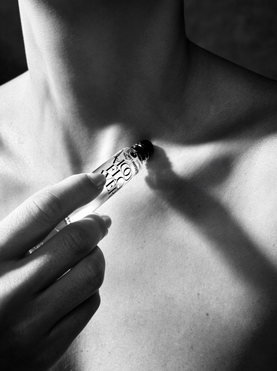 A close-up black and white photograph of a hand holding a clear glass rollerball perfume bottle against a person's neck and upper chest.