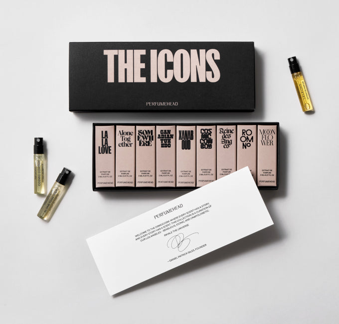 A perfume gift set  titled 'THE ICONS' arranged on a light gray background. The set includes a black presentation box with large beige lettering, a row of nine pale pink perfume bottles, three small gold perfume vials, and a white printed card with text and a signature.