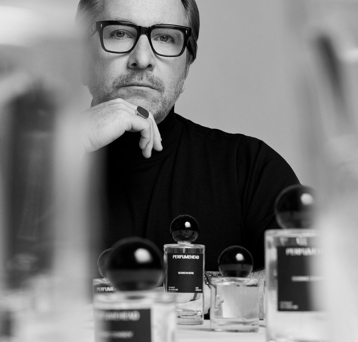 A black and white editorial photograph of a man with glasses and a beard, wearing a black turtleneck and resting his chin on his hand. In the foreground, several perfume bottles with black spherical caps are arranged on a table.