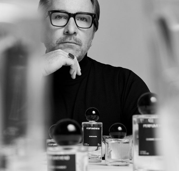 A black and white editorial photograph of a man with glasses and a beard, wearing a black turtleneck and resting his chin on his hand. In the foreground, several perfume bottles with black spherical caps are arranged on a table.