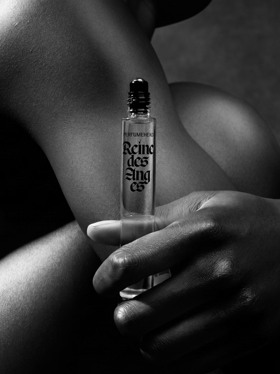 A black and white close-up of a hand holding a clear glass perfume rollerball bottle labeled 'Reine des Anges' against a dark, moody background.