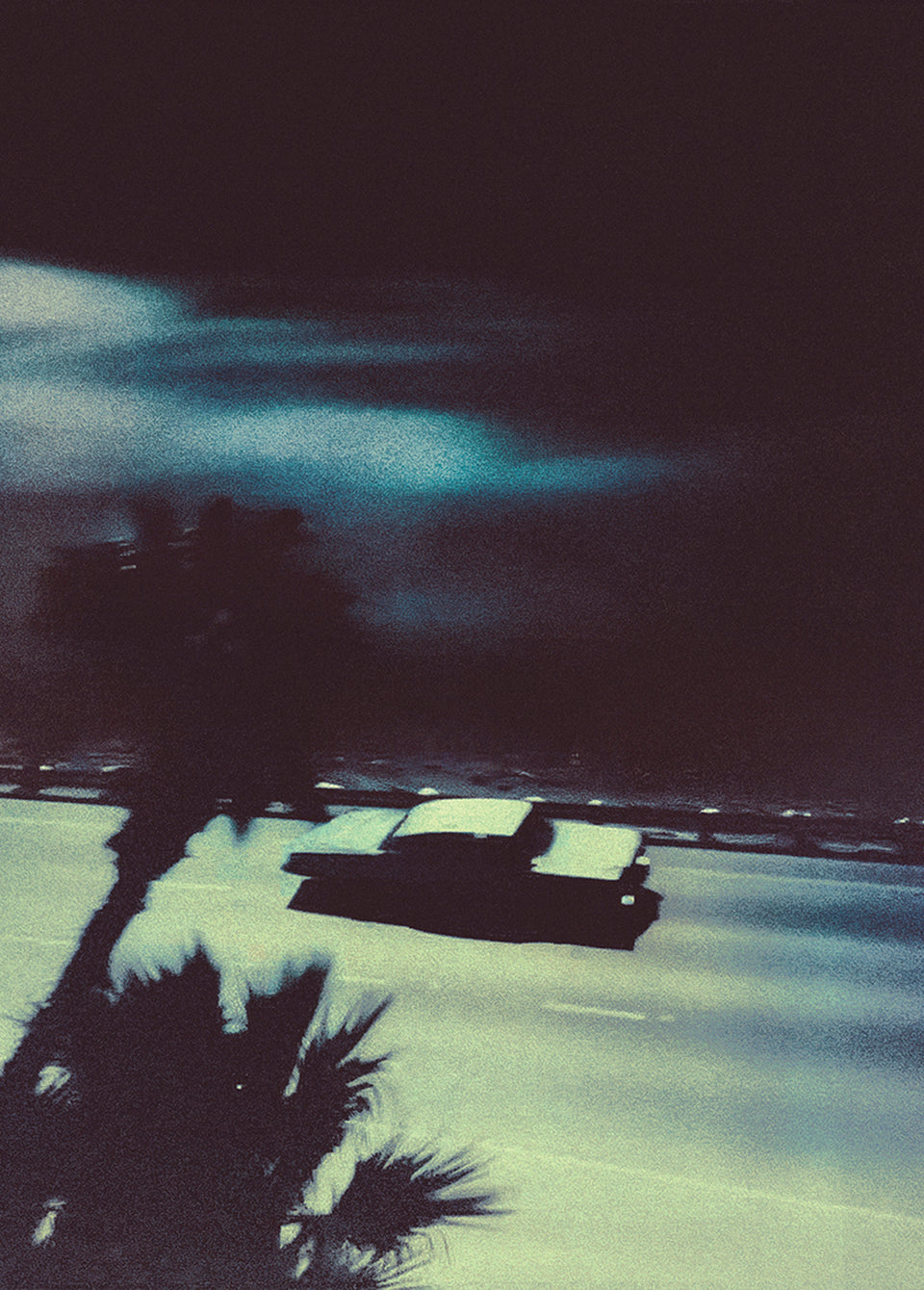 A moody, grainy photograph of a vintage car driving along a coastal road at night, with palm tree silhouettes in the foreground and a dark, dramatic sky.