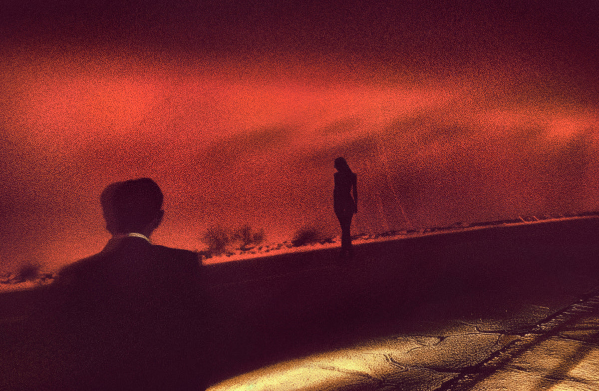 A dramatic, grainy art photograph featuring silhouettes of a man in a suit and a woman standing against a fiery red and orange sky, with a bright light reflecting on the ground.