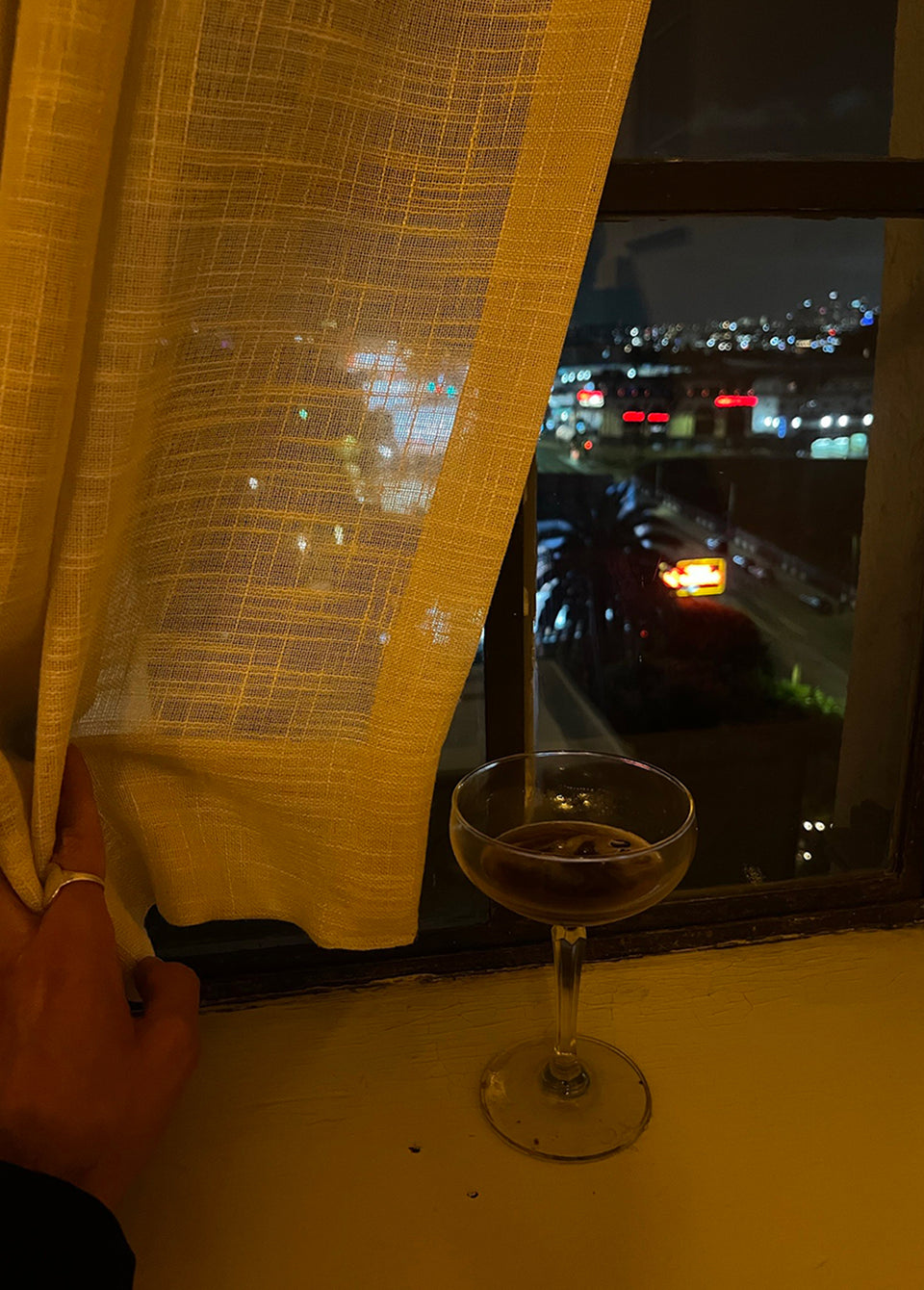 A hand pulls back a sheer yellow curtain next to a window, revealing a nighttime city street scene outside. A coupe glass with a dark drink sits on the windowsill.