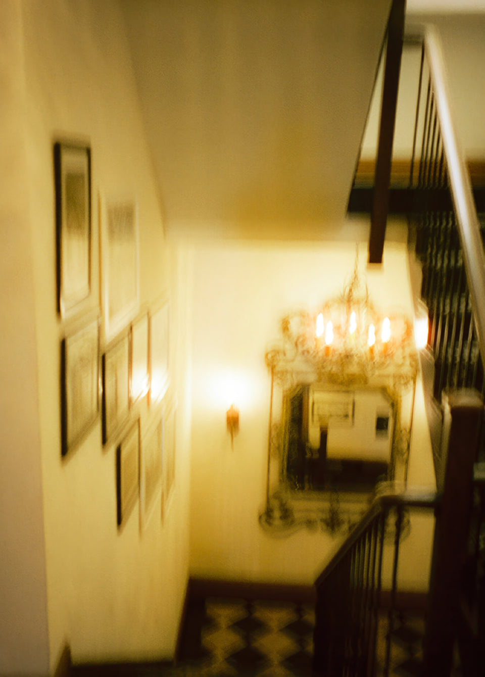 A warm, dimly lit interior staircase with framed wall art on the left, a decorative mirror, a chandelier, and dark wooden railings.