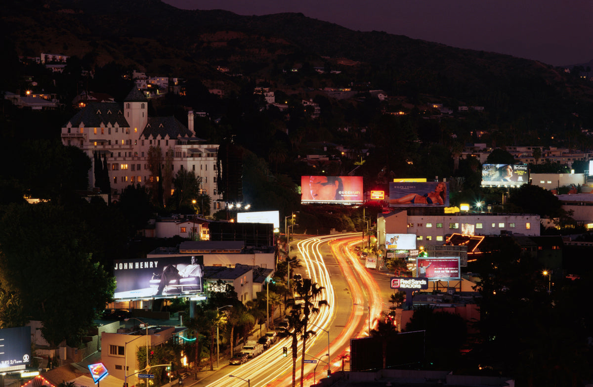 Nighttime aerial view of a city street with long light trails from moving vehicles, surrounded by buildings, illuminated billboards, and dark hills in the background.