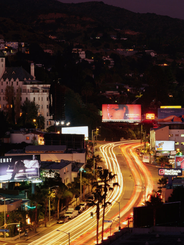 Nighttime city street scene with a curved highway showing long light trails from moving vehicles, surrounded by buildings, palm trees, and large illuminated billboards on a dark hillside.