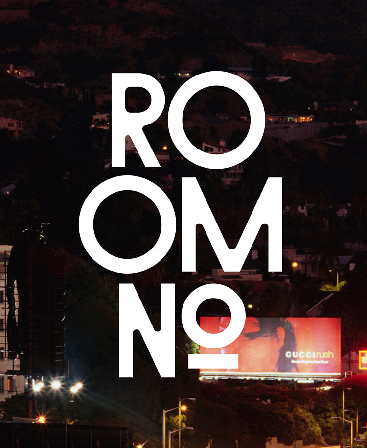 A nighttime city scene with large white stacked text reading 'ROOOM NO' over a dark hillside neighborhood, plus a billboard and streetlights in the background.