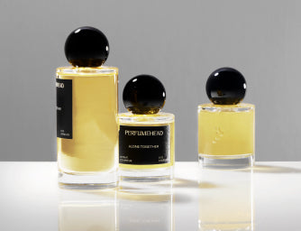 Three clear glass perfume bottles with black spherical caps and pale yellow liquid, arranged on a white reflective surface against a light gray studio background. The center bottle label reads 'PERFUMEHEAD'.