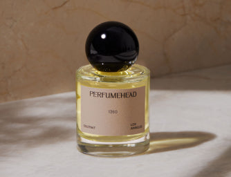 A bottle of Perfumehead 1360 coltrait with a black spherical cap and a beige label, set against a textured beige background with soft shadows.
