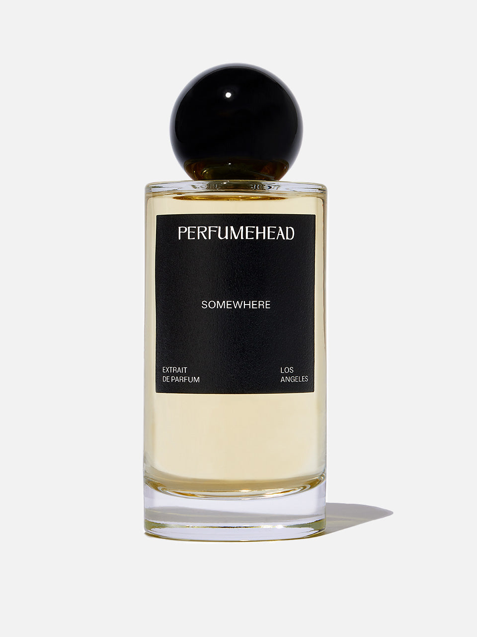 A bottle of Perfumehead 'SOMEWHERE' extrait de parfum, featuring a clear glass bottle with pale yellow liquid, a black label, and a spherical black cap, set against a light gray background.