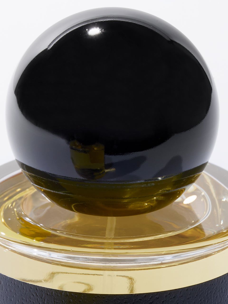 A close-up of a glossy black spherical bottle top resting on a clear glass base with a gold liquid, set against a plain light background.