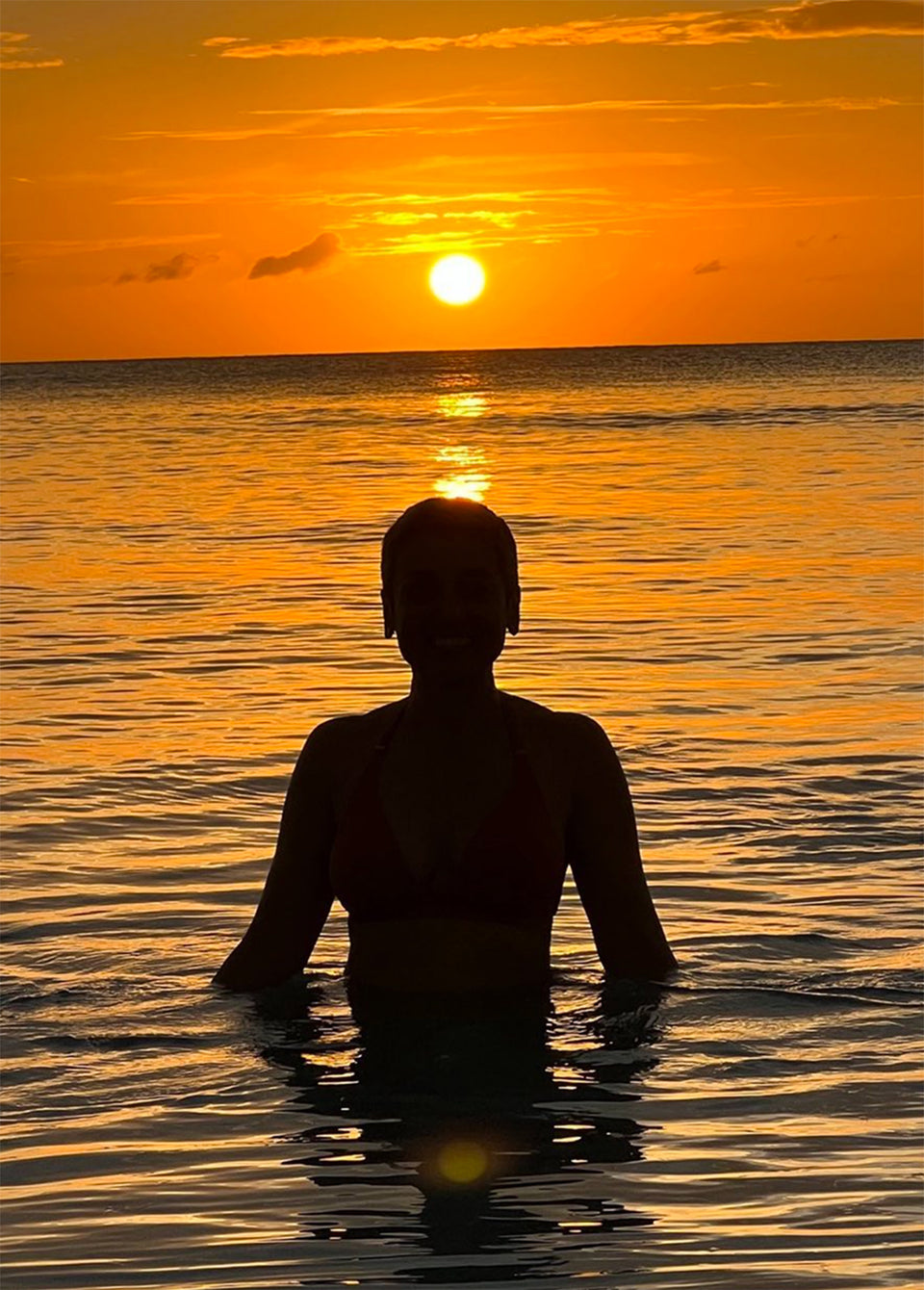 A person standing in ocean water at sunset, shown in silhouette against a glowing orange sky and reflective sea.