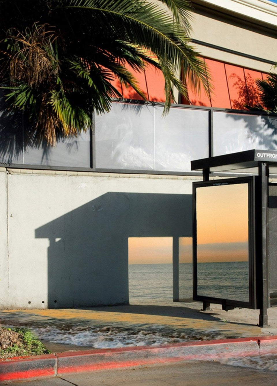 A modern outdoor bus stop shelter beside a building with large windows and palm tree shadows, showing an ocean sunset scene in the shelter panel and reflected in the window.